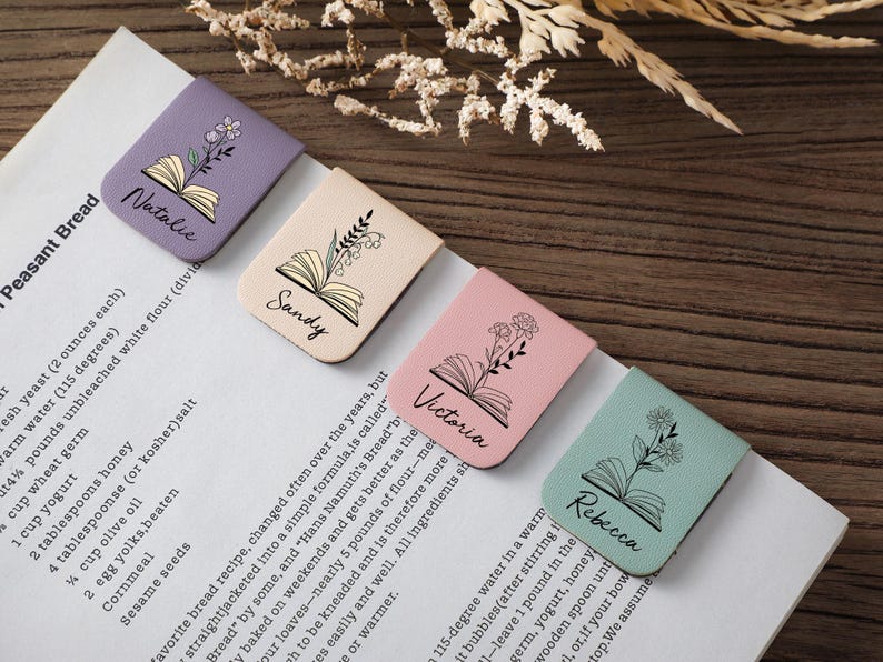 Puede incluir: Cuatro marcadores de libros de cuero personalizados en colores pastel: lavanda, beige, rosa y verde menta. Cada uno presenta un dise&ntilde;o floral, una ilustraci&oacute;n de libro abierto y un nombre: Natalie, Sandy, Victoria y Rebecca. Los marcadores est&aacute;n en una p&aacute;gina blanca.