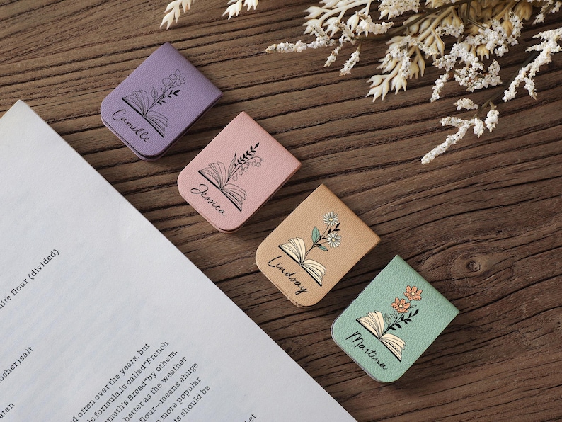 Puede incluir: Cuatro marcadores de libros personalizados de cuero en colores pastel: lavanda, rosa, beige y verde menta. Cada uno presenta una ilustraci&oacute;n de libro abierto con un dise&ntilde;o floral y un nombre: Camille, Jessica, Lindsay y Martina. Sobre una superficie de madera.