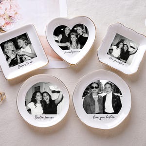 May include: A collection of white ceramic trinket dishes in various shapes, each featuring a black and white photograph and gold trim. Text includes phrases like "Cheers to us" and "Love you bestie."