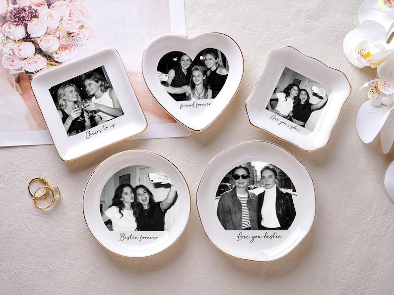 May include: A collection of white ceramic trinket dishes in various shapes, each featuring a black and white photograph and gold trim. Text includes phrases like "Cheers to us" and "Love you bestie."
