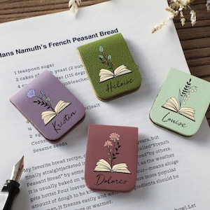 Puede incluir: Cuatro marcadores de libros de cuero con forma de libro en tonos morado, verde y rosa, cada uno con un dise&ntilde;o floral y un nombre: Kristin, Heloise, Louise y Dolores. Los marcadores est&aacute;n en una p&aacute;gina de un libro de cocina.