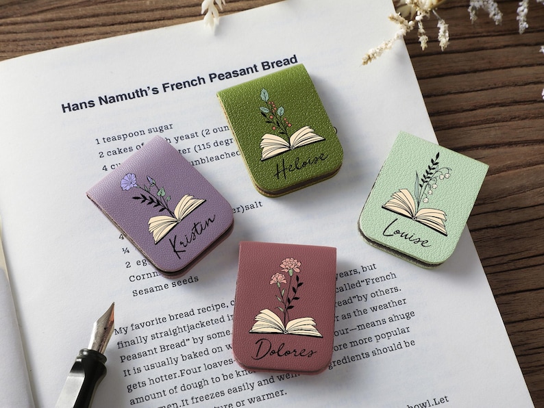 Puede incluir: Cuatro marcadores de libros de cuero con forma de libro en tonos morado, verde y rosa, cada uno con un dise&ntilde;o floral y un nombre: Kristin, Heloise, Louise y Dolores. Los marcadores est&aacute;n en una p&aacute;gina de un libro de cocina.