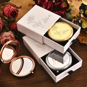 Könnte beinhalten: Kompaktspiegel in weißen Schachteln mit dem Namen "Astrid" und einem Rosendesign. Ein Spiegel ist geöffnet, mit einem roségoldenen Rahmen. Die anderen Spiegel sind gold- und silberfarben. Rosen sind im Hintergrund.