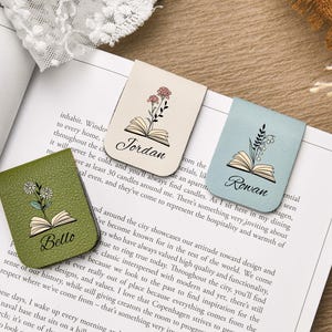 Puede incluir: Tres marcadores de cuero en beige, verde salvia y azul claro, cada uno con un dise&ntilde;o floral y un nombre. Los marcadores est&aacute;n en un libro, con los nombres "Jordan", "Bello" y "Rowan" en escritura cursiva.