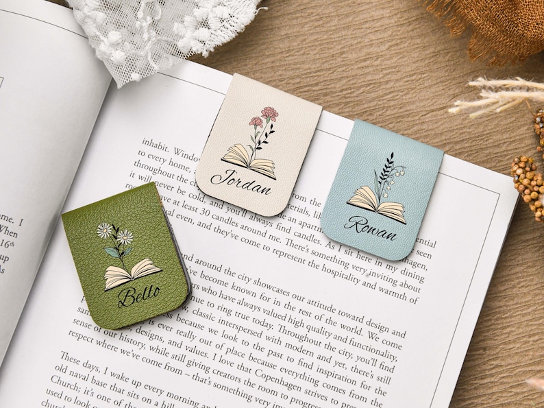 Puede incluir: Tres marcadores de cuero en beige, verde salvia y azul claro, cada uno con un dise&ntilde;o floral y un nombre. Los marcadores est&aacute;n en un libro, con los nombres "Jordan", "Bello" y "Rowan" en escritura cursiva.