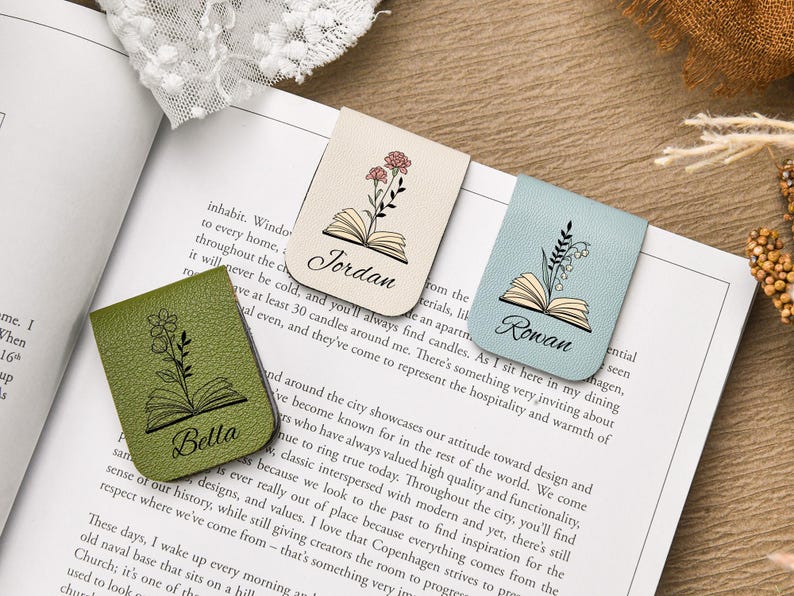 Puede incluir: Tres marcadores de cuero con dise&ntilde;os florales y nombres: Bella, Jordan y Rowan. Los marcadores son verdes, beige y azules, cada uno con una ilustraci&oacute;n floral diferente y un dise&ntilde;o de libro abierto. Est&aacute;n colocados en un libro abierto.