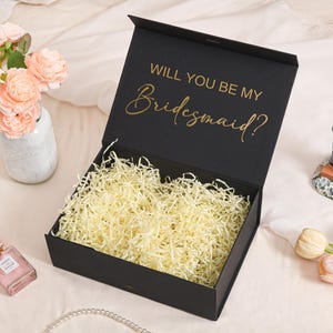 以下が含まれることがあります： 「WILL YOU BE MY Bridesmaid?」の金色の文字が入った黒いギフトボックス。ボックスは開いており、黄色い紙吹雪が詰められています。ウェディングケーキのトッパーと香水瓶も確認できます。