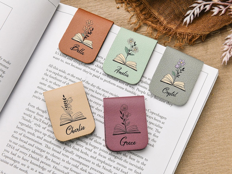 Puede incluir: Cinco marcadores magn&eacute;ticos de cuero en varios colores, cada uno con un dise&ntilde;o floral y un nombre: Bella, Amelia, Crystal, Charlie y Grace. Los marcadores est&aacute;n sobre un libro abierto.