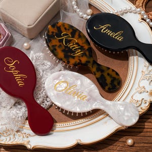 May include: Four personalized hairbrushes in a decorative arrangement. The brushes are in various colors: burgundy, black, tortoiseshell, and pearlescent white. Each brush has a name in gold script: Sophia, Amelia, Kitty, and Olivia. The brushes are on a white and gold ornate plate.