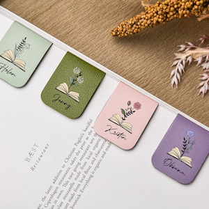 Puede incluir: Cuatro marcadores de cuero en colores pastel: verde menta, verde oliva, rosa y lavanda. Cada uno presenta un dise&ntilde;o de libro abierto con una ilustraci&oacute;n floral y un nombre: Helen, Jenny, Kristin y Oksana. Los marcadores est&aacute;n en una p&aacute;gina blanca.