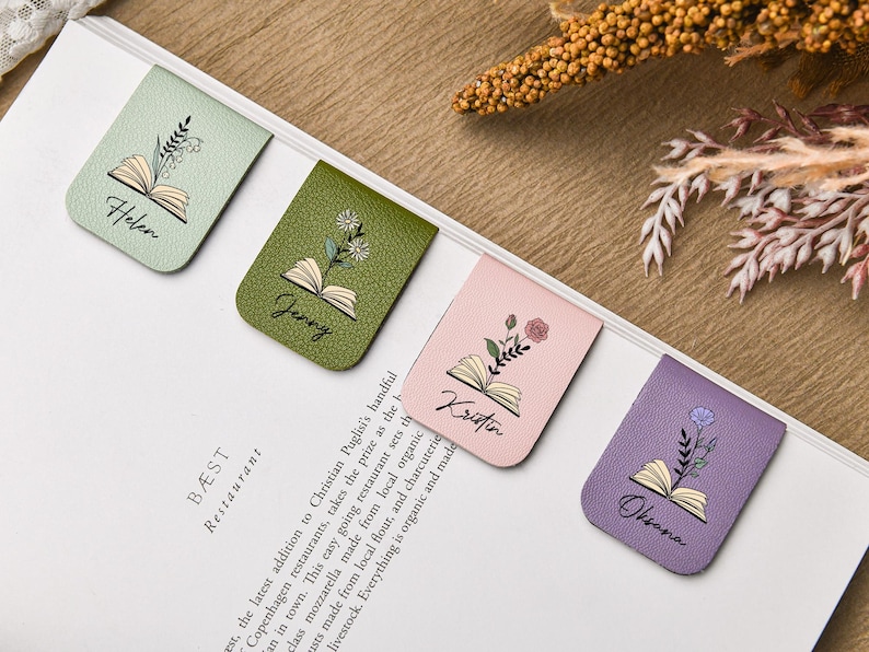 Puede incluir: Cuatro marcadores de cuero en colores pastel: verde menta, verde oliva, rosa y lavanda. Cada uno presenta un dise&ntilde;o de libro abierto con una ilustraci&oacute;n floral y un nombre: Helen, Jenny, Kristin y Oksana. Los marcadores est&aacute;n en una p&aacute;gina blanca.