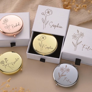 May include: A collection of personalized compact mirrors in various metallic finishes, including rose gold, gold, and silver. Each mirror is engraved with a name and floral design, presented in a white gift box with a black ribbon pull.