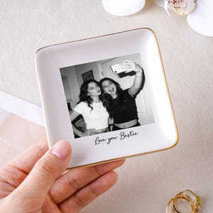 May include: A white ceramic trinket dish with a gold rim, featuring a black and white photo of two women taking a selfie. The words "Love you Bestie" are written below the photo. The dish is held in a hand.