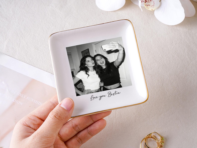 May include: A white ceramic trinket dish with a gold rim, featuring a black and white photo of two women taking a selfie. The words "Love you Bestie" are written below the photo. The dish is held in a hand.