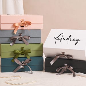 Può includere: Una pila di scatole regalo in vari colori, tra cui rosa, grigio, verde, verde acqua, bianco e nero. Ogni scatola ha un nastro e un fiocco abbinati. La scatola bianca superiore ha il nome "Audrey" in corsivo nero.