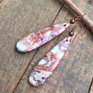 May include: A pair of teardrop-shaped earrings with a marbled pattern in shades of white, orange, and brown. The earrings have copper-colored ear wires and small beads. The earrings are displayed on a wooden surface.