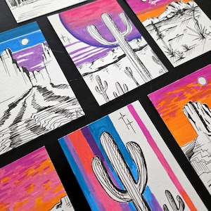 May include: A collection of rectangular art cards featuring ink and marker illustrations of desert landscapes. The drawings depict saguaro cacti, rock formations, and sunsets in vibrant shades of pink, orange, purple, and blue, with one card showing the text Miles to go.