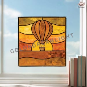 May include: Stained glass window art featuring a hot air balloon design in shades of orange, yellow, and brown. The artwork depicts a sunset scene with the text "COLOR DREAMLIGHT". The window is square and framed in black.