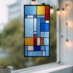 以下が含まれることがあります： 黒いフレームの長方形ステンドグラスの窓。デザインは、青、黄、赤、白の色合いの幾何学的な形を特徴としています。「COLORDREAMLIGHT」の文字がガラスに表示されています。窓は、電飾の付いた窓の前に吊るされています。