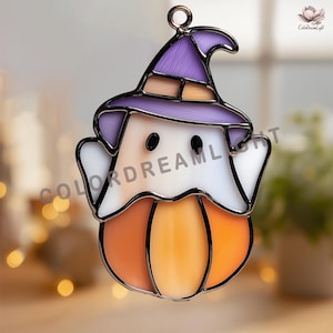 Op de afbeelding: Een glas-in-lood suncatcher in de vorm van een geest met een paarse heksenhoed, zittend op een oranje pompoen. Het object heeft zwarte metalen soldeerlijnen, twee zwarte ronde ogen en een klein metalen oogje aan de bovenkant om op te hangen.