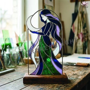 Op de afbeelding: Een gebrandschilderde glazen sculptuur van een vrouw in een vloeiende jurk, die een halve maan vasthoudt. Het kunstwerk heeft tinten blauw, groen en paars, tegen een witte achtergrond. Het is gemonteerd op een donkere houten voet.