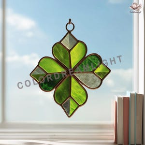 May include: A stained glass four-leaf clover suncatcher with shades of green and white, framed in a copper-colored metal. The clover hangs from a small ring, with the text "COLORDREAMLIGHT" visible. The background is a blue sky.