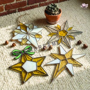 May include: Four stained glass ornaments in star and flower shapes, featuring yellow and white glass panels with dark metal soldering. One star ornament includes a green glass bow accent. A small potted cactus sits above the ornaments.