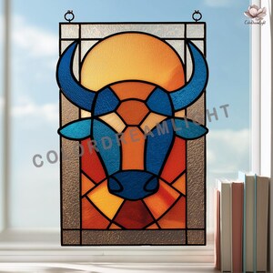 Op de afbeelding: Gebrandschilderd glaspaneel met een gestileerde stierenkop met blauwe hoorns en een geometrisch ontwerp in oranje, rood en blauw. Het paneel heeft een zonachtige vorm op de achtergrond en is omlijst door een donkere rand. Het paneel hangt voor een raam.