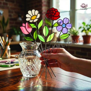 Op de afbeelding: Een verzameling kleurrijke glas-in-lood bloemstelen, waaronder een madeliefje, roos, tulp en andere bloemen. De bloemen worden voor een heldere glazen vaas gehouden. De bloemen hebben groene bladeren en metalen stelen.