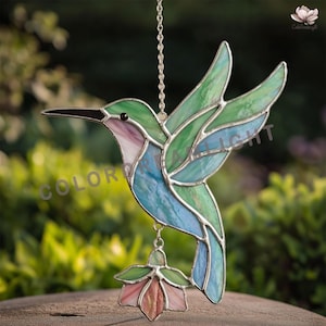 Puede incluir: Atrapasoles de colibrí de vidrieras en tonos verdes, azules y morados. El pájaro está suspendido de una cadena y presenta un colgante de flores. Las palabras "COLOR LIGHT" son visibles.