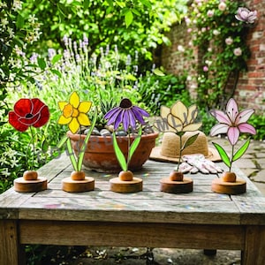 May include: Five colorful stained glass flower sculptures on wooden bases. The flowers include a red poppy, yellow daffodil, purple coneflower, cream-colored lotus, and pink lily. They are arranged on a weathered wooden table in a garden setting.