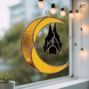 May include: A stained glass suncatcher featuring a yellow crescent moon with a textured finish. A black bat hangs upside down within the moon's curve. The suncatcher is suspended in front of a window, with string lights in the background.