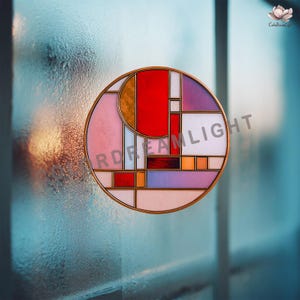 Puede incluir: Una ventana redonda de vidriera con un diseño geométrico. Tiene un marco dorado y un patrón abstracto colorido con formas rojas, rosas, moradas y naranjas. El texto "RDREAMLIGHT" es visible.