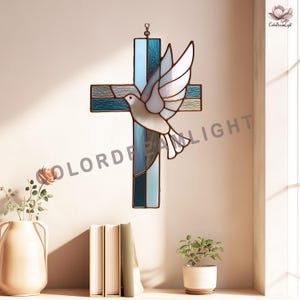 May include: Stained glass cross with a dove design. The cross features shades of blue and green glass, with a white dove in flight. The artwork is designed to hang and is displayed against a neutral wall. The dove has outstretched wings.