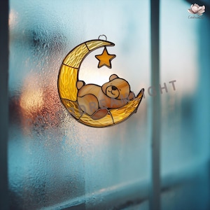 May include: Stained glass suncatcher featuring a sleeping teddy bear on a yellow crescent moon with a hanging star. The bear is light brown, and the moon and star are yellow. The suncatcher is hanging in front of a window.