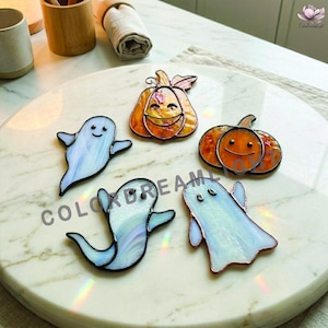 May include: A collection of four stained glass ornaments featuring two pumpkins with orange textured glass and two ghosts with iridescent white glass. Each piece is outlined in dark metal solder, showcasing smiling faces and simple, rounded shapes.