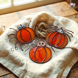 May include: Three stained glass spider ornaments with orange pumpkin-shaped bodies. Each spider features dark metal wire legs and a small head with either googly eyes or a simple hand-drawn face. The text ColorDreamLight is visible in the image.