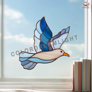 May include: Stained glass bird in flight with shades of blue and white. The bird has a tan body and an orange beak. The wings are a mosaic of blue hues, and the bird is set against a blue sky background. The text "COLORDREAMLIGHT" is visible.