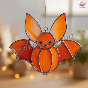 May include: A stained glass bat suncatcher in shades of orange and amber with dark metal soldering. The bat features large pointed ears, two small black eyes, and a small triangular nose. It hangs from a thin cord for display in a window.