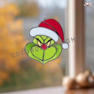 May include: A stained glass ornament featuring the Grinch's face, wearing a red and white Santa hat. The Grinch has a green face, yellow eyes, and a red nose. The ornament has the text "COLOR DREAM LIGHT".