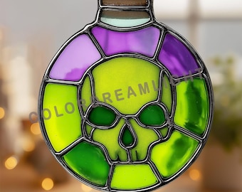 Puede incluir: Un atrapasoles de vidrieras con forma de frasco de poción redondo con un diseño de calavera en el centro. La calavera es de color verde lima, rodeada por segmentos de vidrio morado y verde, unidos por líneas de plomo de metal oscuro.