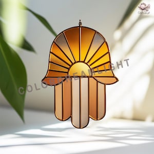 May include: A stained glass Hamsa hand suncatcher with a sunburst design in gold, orange, and beige. The hand-shaped ornament has a copper-coloured metal frame and is designed to hang. The text "COLOR DREAM LIGHT" is visible.