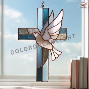 May include: A stained glass cross suncatcher featuring a white dove in flight. The cross is constructed from various shades of blue and clear textured glass panels held together by copper-colored metal cames. The text COLORDREAMLIGHT is visible.