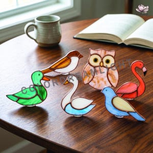 May include: A collection of colorful stained glass bird figurines, including a duck, swan, owl, flamingo, and two other birds. The figurines are displayed on a wooden table near a window and a book.