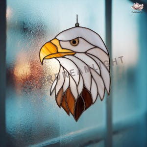 May include: Stained glass eagle head suncatcher with a detailed design. The eagle features a yellow beak, brown and white feathers, and a focused gaze. The suncatcher is suspended by a chain, perfect for window or wall decor.