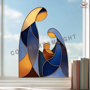 May include: A stained glass artwork depicting the Holy Family. The image shows a stylized representation of Mary, Joseph, and baby Jesus, with a colour palette of blues, gold, and brown. The piece is set against a bright blue sky.