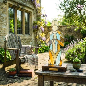 May include: A stained glass figure of a person kneeling with hands clasped in prayer. The figure has a geometric design with yellow, blue, and orange hues. It is mounted on a dark wooden base and placed on a wooden table outdoors.