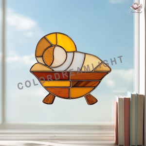 May include: A stained glass window decoration featuring a ram design. The artwork uses a variety of colors, including shades of yellow, brown, and white, and is set against a blue sky background. The piece is displayed on a window.