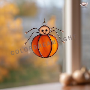 May include: A stained glass spider decoration with an orange body and a smiling face. The spider has thin, dark legs and a small loop for hanging. The background is blurred, with the text "COLOR DREAM LIGHT" visible.