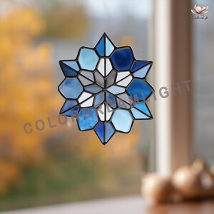 Può includere: Decorazione per finestra in vetro colorato a forma di stella. Il design presenta una varietà di forme geometriche blu, bianche e grigie, incorniciate da un bordo in metallo scuro. Le parole "COLOR DREAM LIGHT" sono visibili.